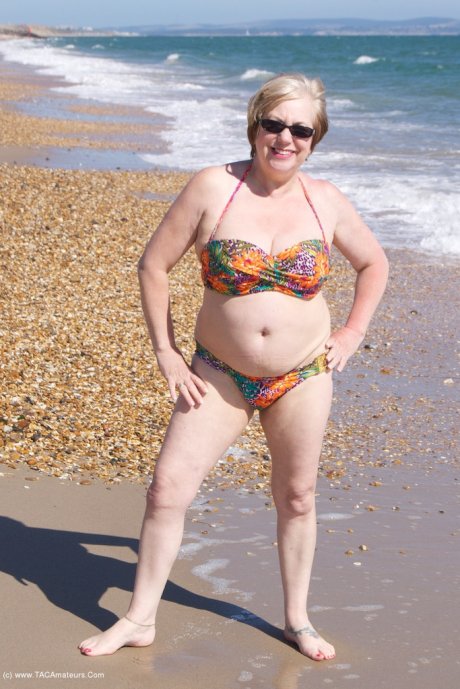 Mature UK woman Speedy Bee wears sunglasses while getting naked at the beach