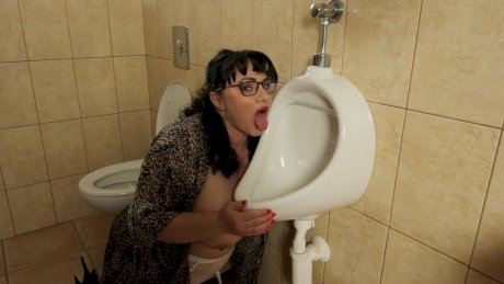 Big titted brunette Mistress Sarah licks a men's urinal while wearing glasses